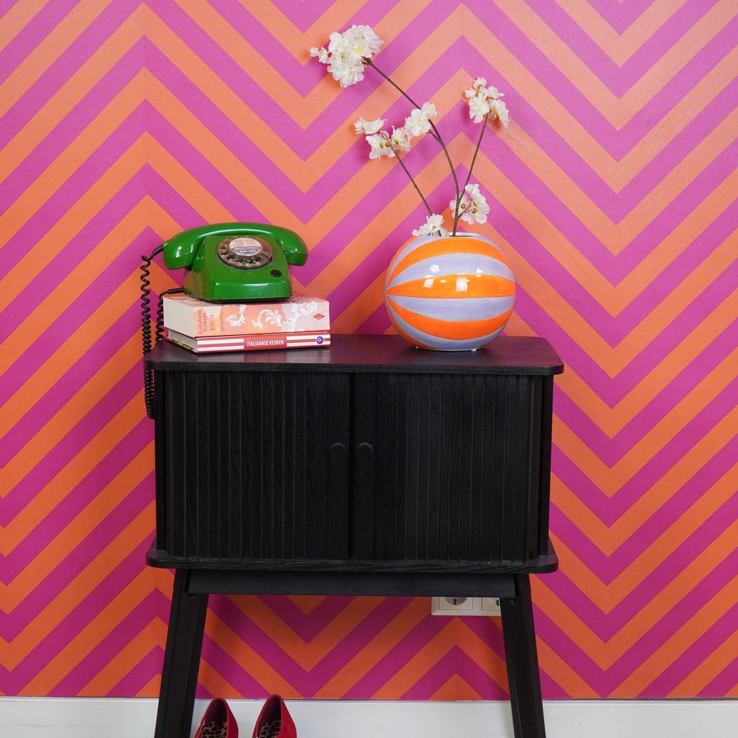 Oranje-lila vaas met witte bloesemtakken op zwarte kast tegen roze-oranje zigzagmuur.